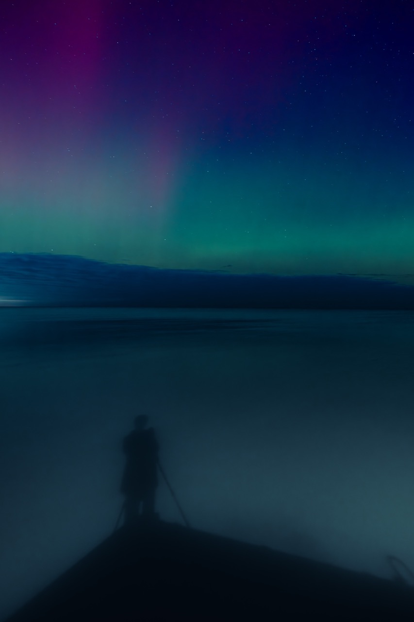 Shadowed self-portrait on a pier beneath auroral glow.