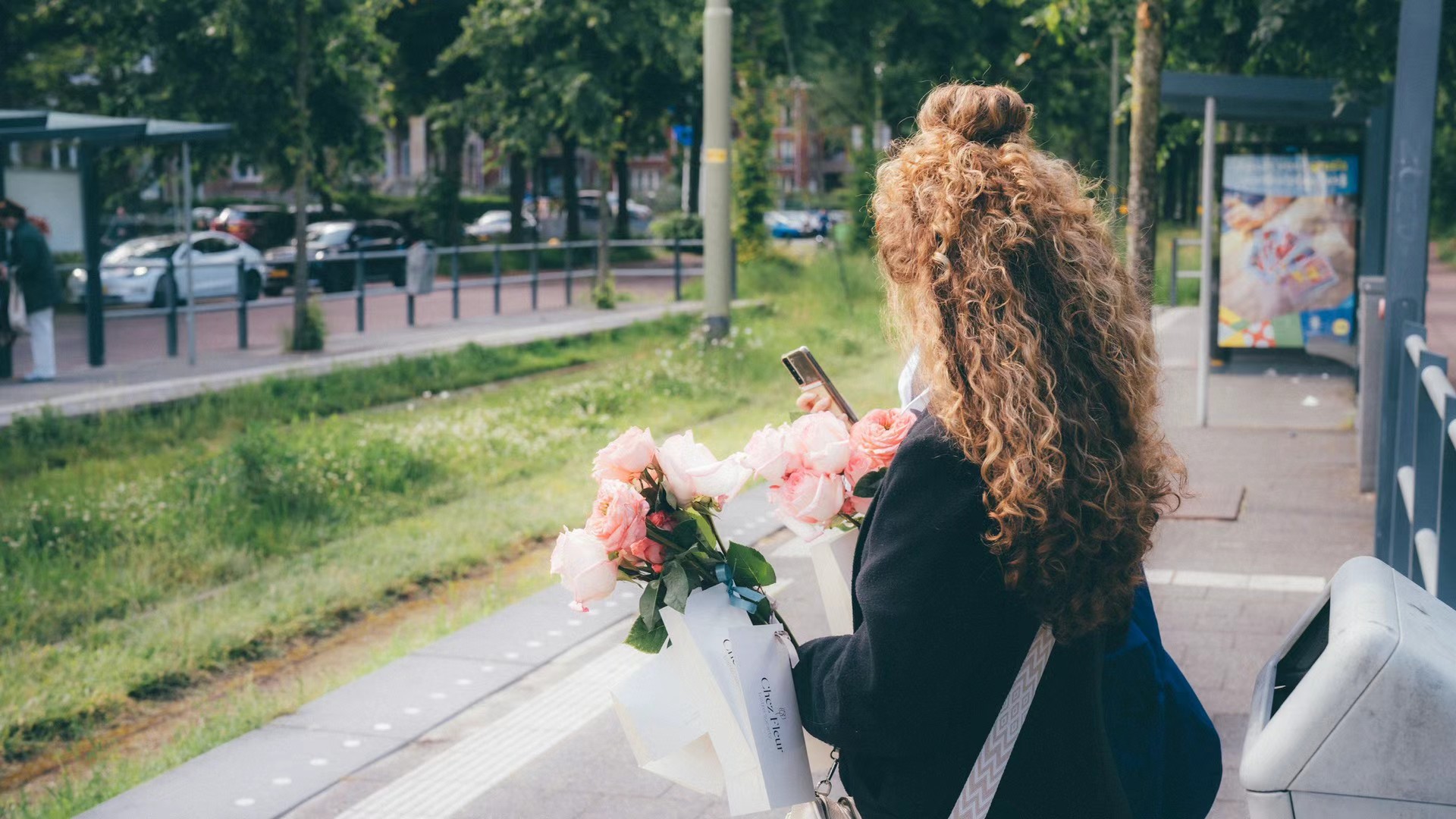 Woman with curly hair carrying blush roses at an Amsterdam tram stop.